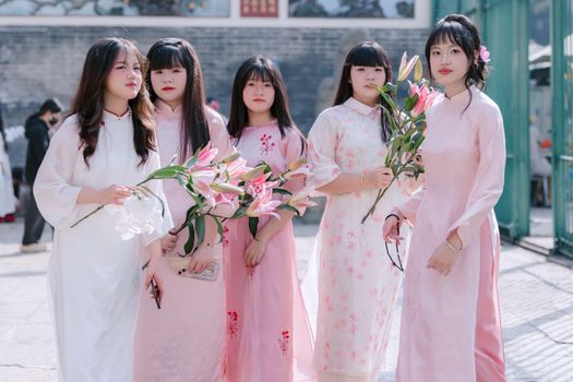 Five young women in traditional dresses holding lilies, outdoors during the day.