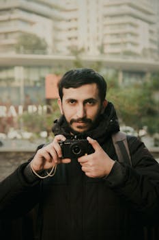 Man in black coat holding a camera with a blurred urban background.