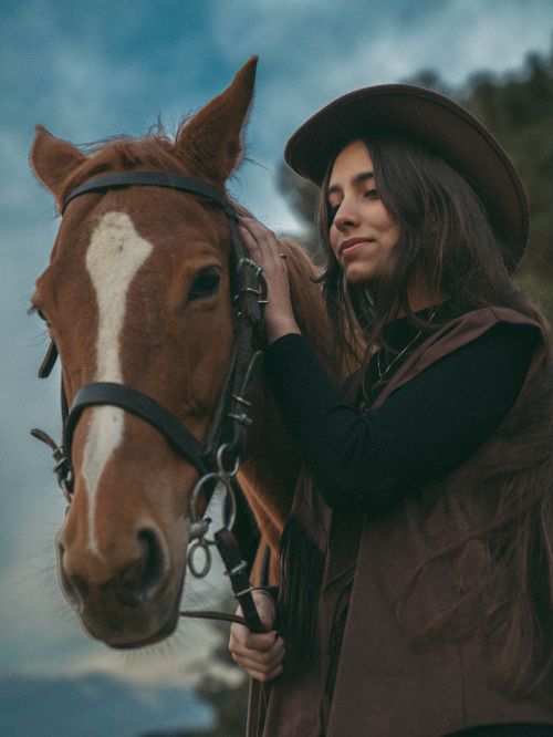 Free Young woman dressed as cowgirl lovingly petting a horse in an outdoor setting at dusk. Stock Photo