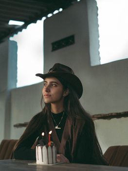 Young woman in a cowboy hat with a cake featuring lit candles, indoors.