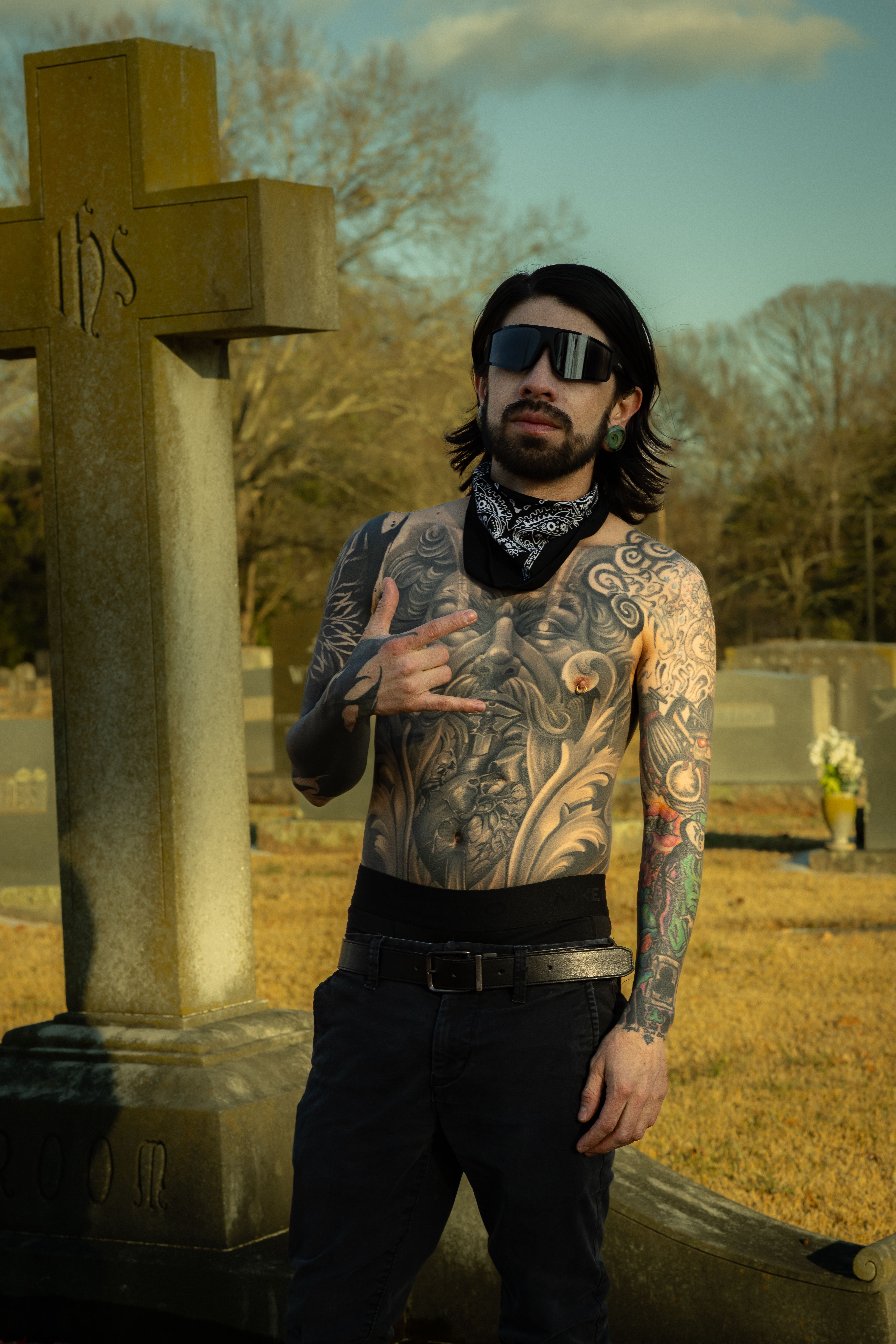 A tattooed man with a bandana poses confidently in a cemetery, showcasing unique body art.