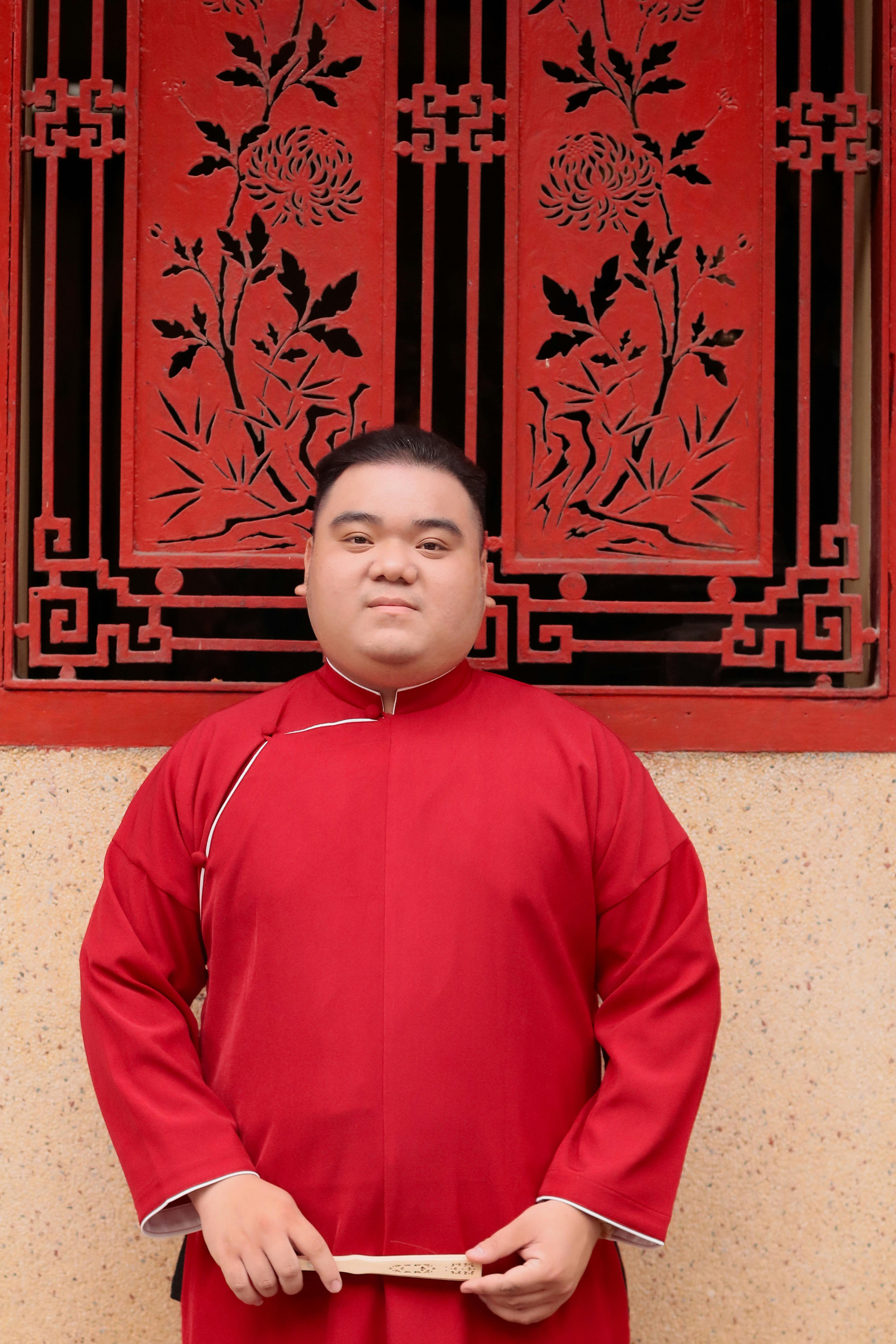 Asian man in red traditional Chinese outfit standing in front of decorative red lattice window.