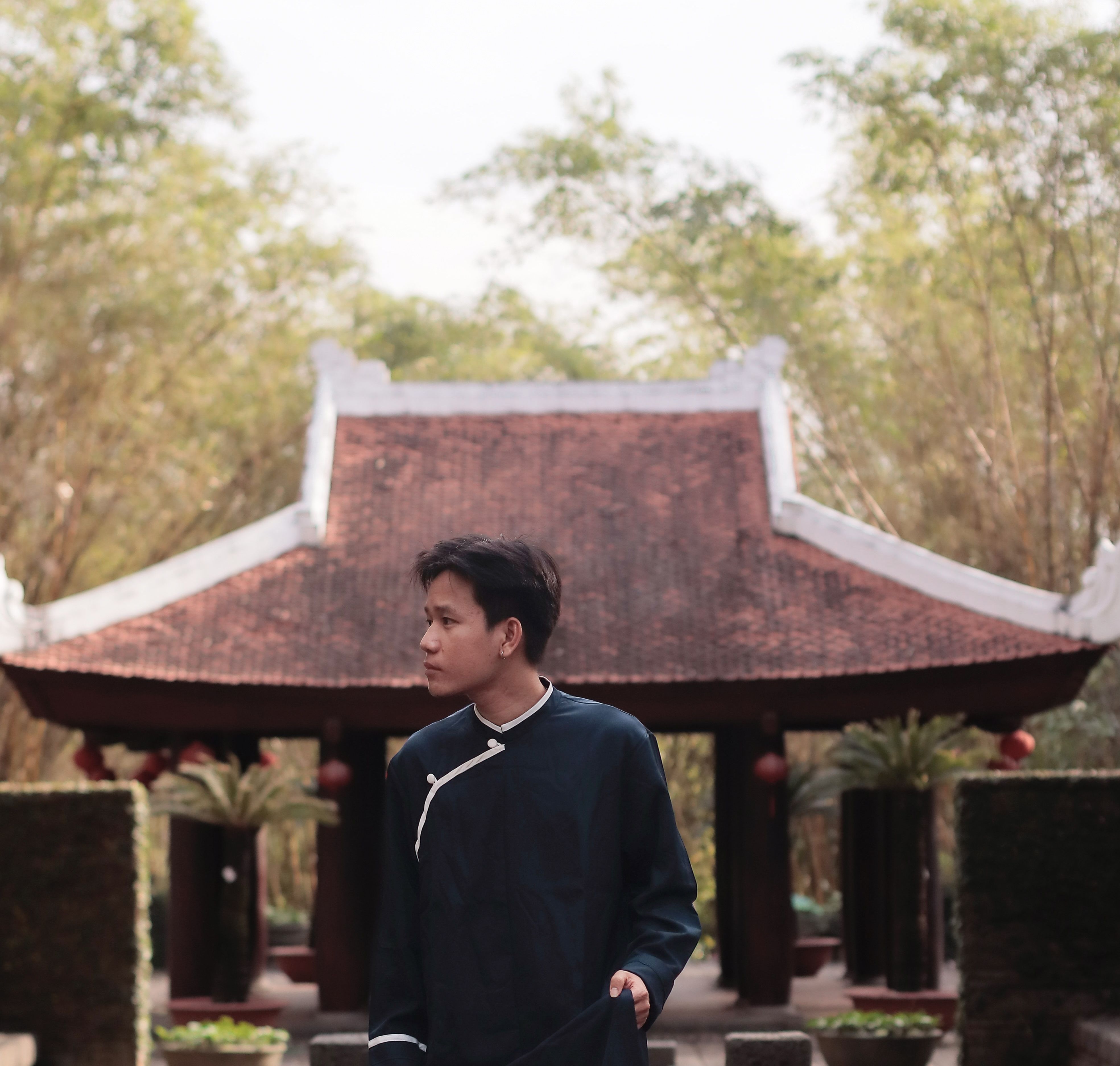 A young man dressed in traditional attire stands in an Asian garden with a pavilion.