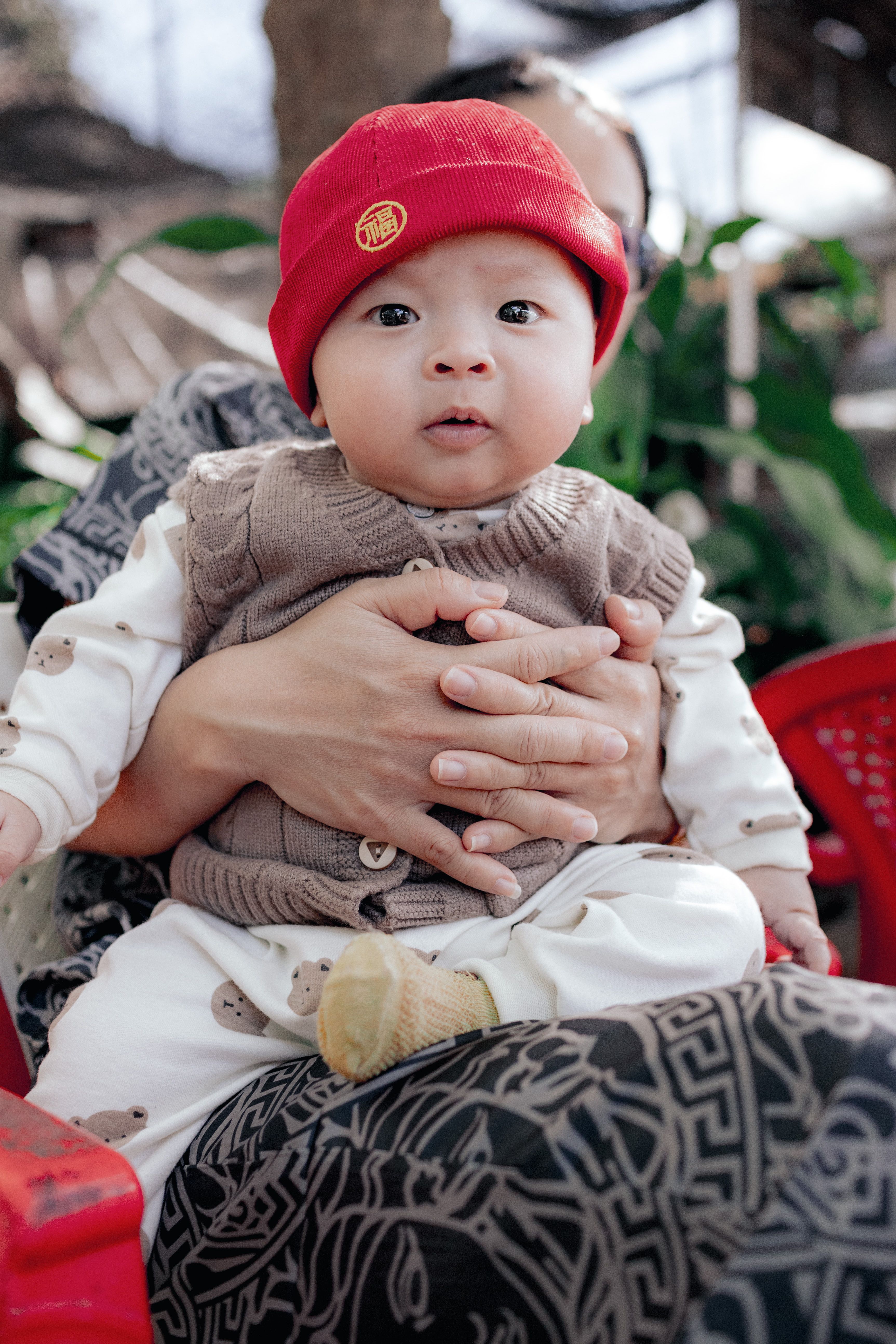 Cute baby wearing a red hat, held by an adult outdoors in Kon Tum, Vietnam.