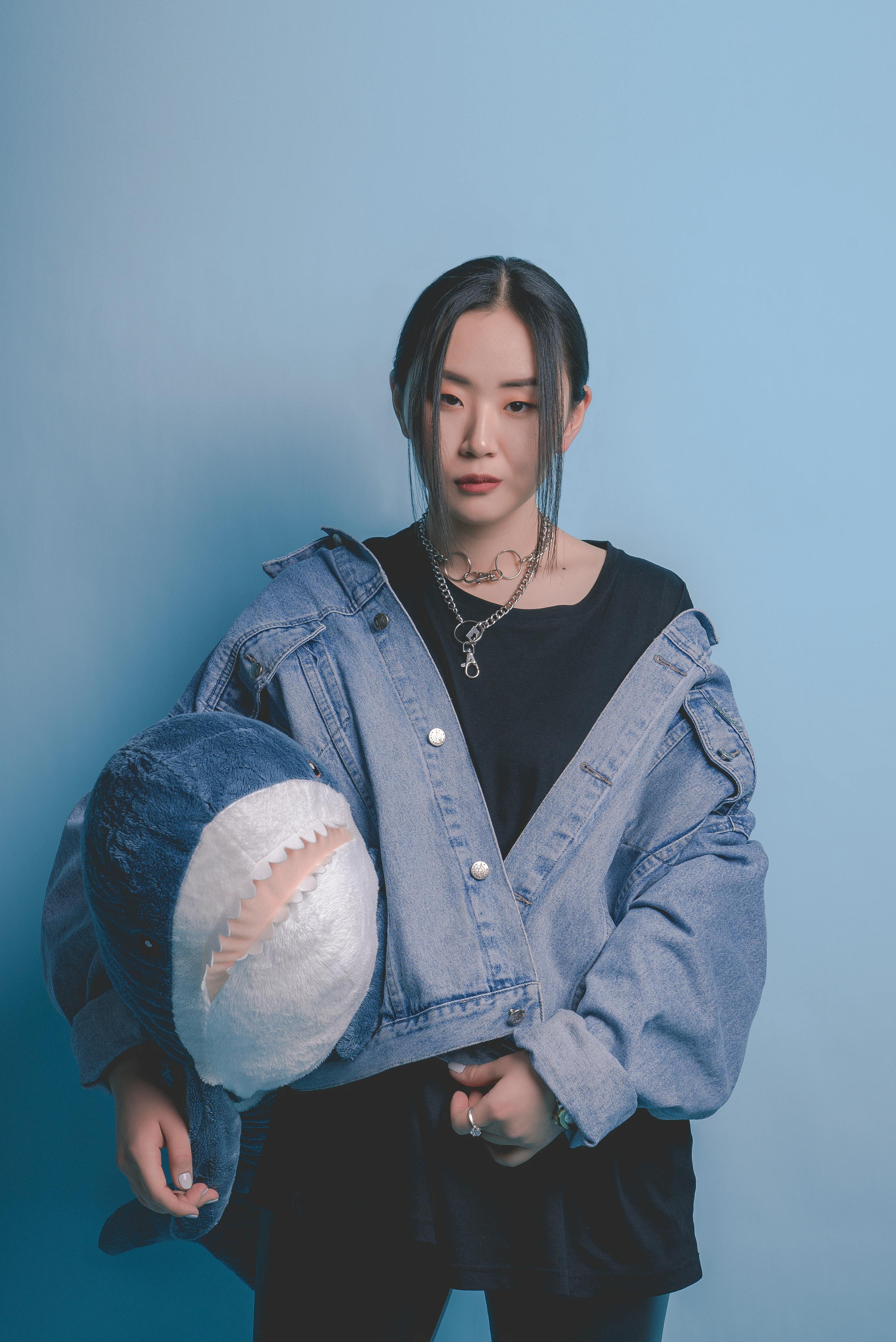 Stylish portrait of an Asian woman in a denim jacket holding a stuffed shark toy against a blue backdrop.