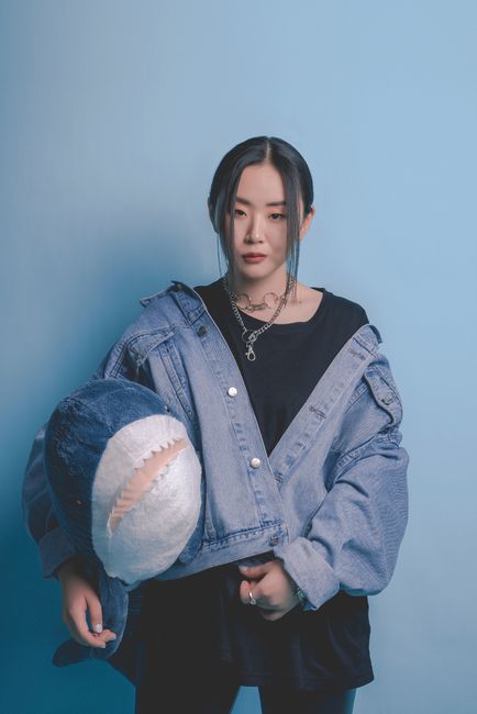 Stylish portrait of an Asian woman in a denim jacket holding a stuffed shark toy against a blue backdrop.