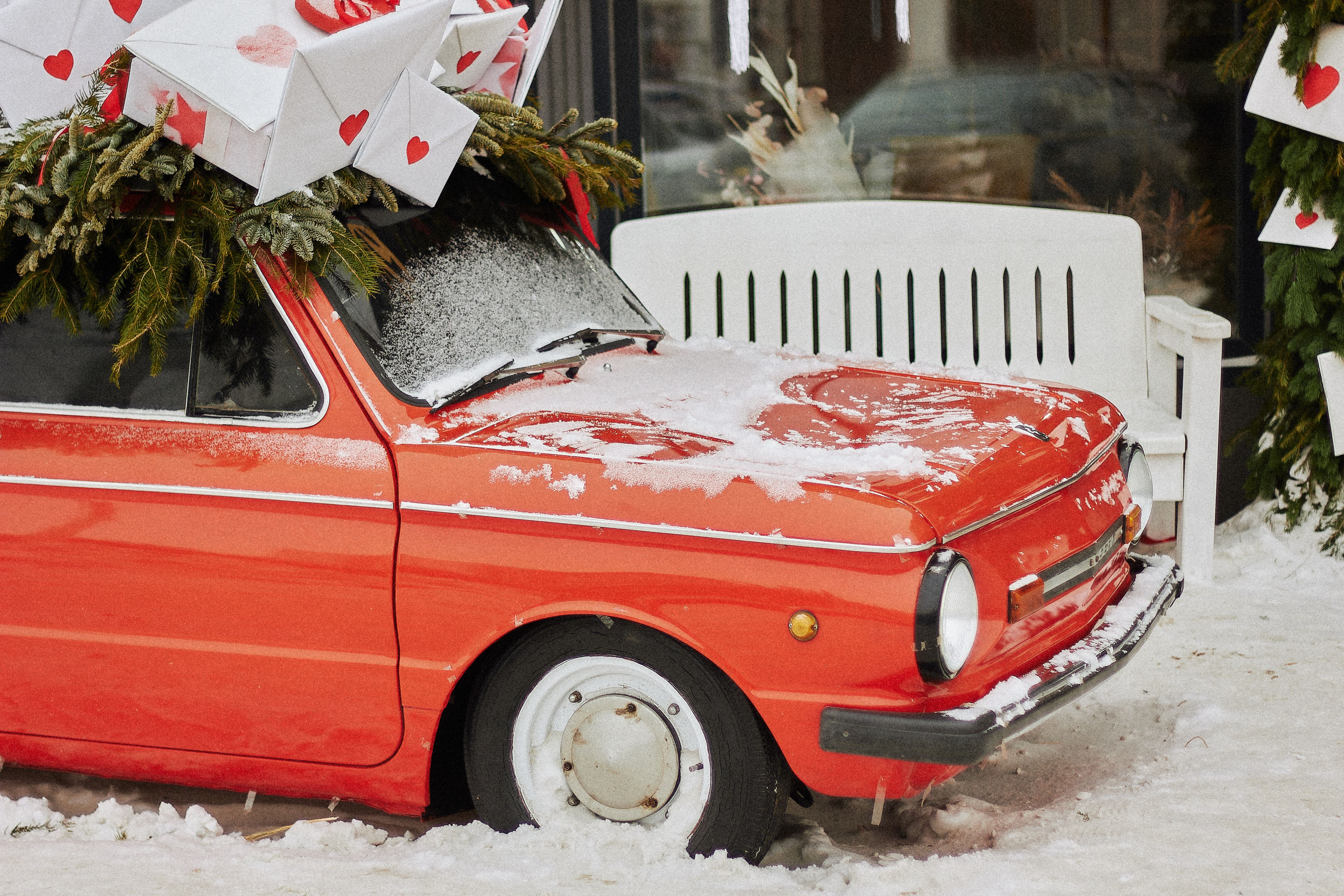 A vintage red car is adorned with holiday letters and pine branches, set in a snowy scene.