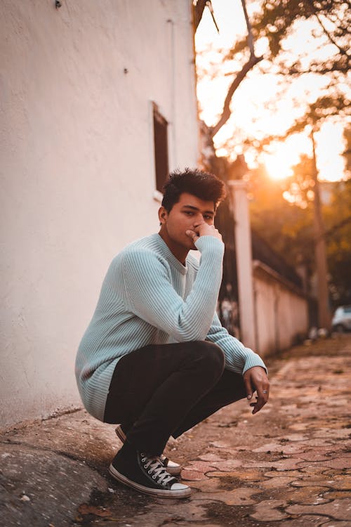 Man in Blue Sweater and Black Pants Sitting on Concrete Wall