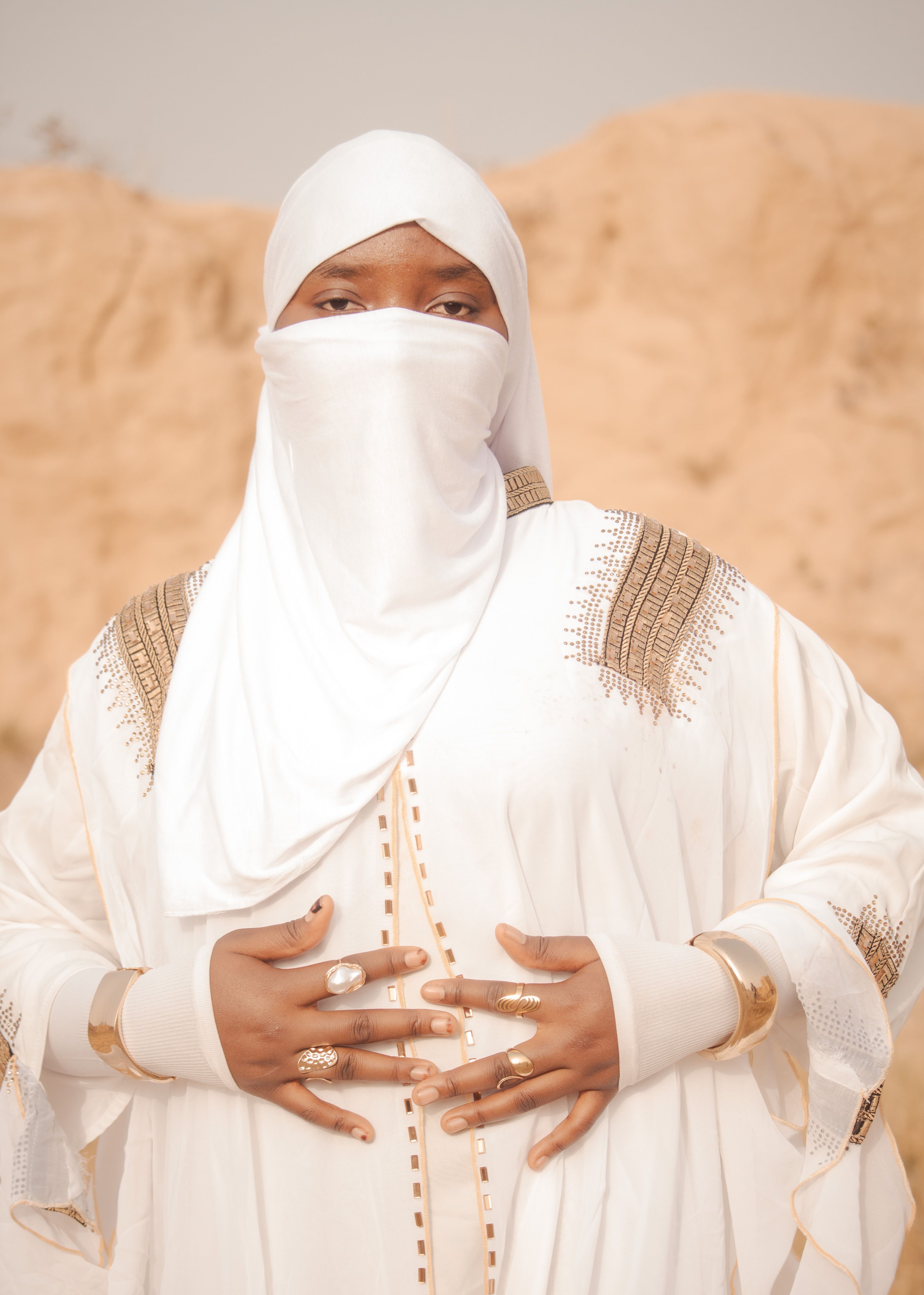 Woman in white traditional attire with veil and jewelry outdoors against a sand dune backdrop.