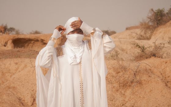 A person in traditional white attire standing in a desert landscape, adjusting their headscarf.