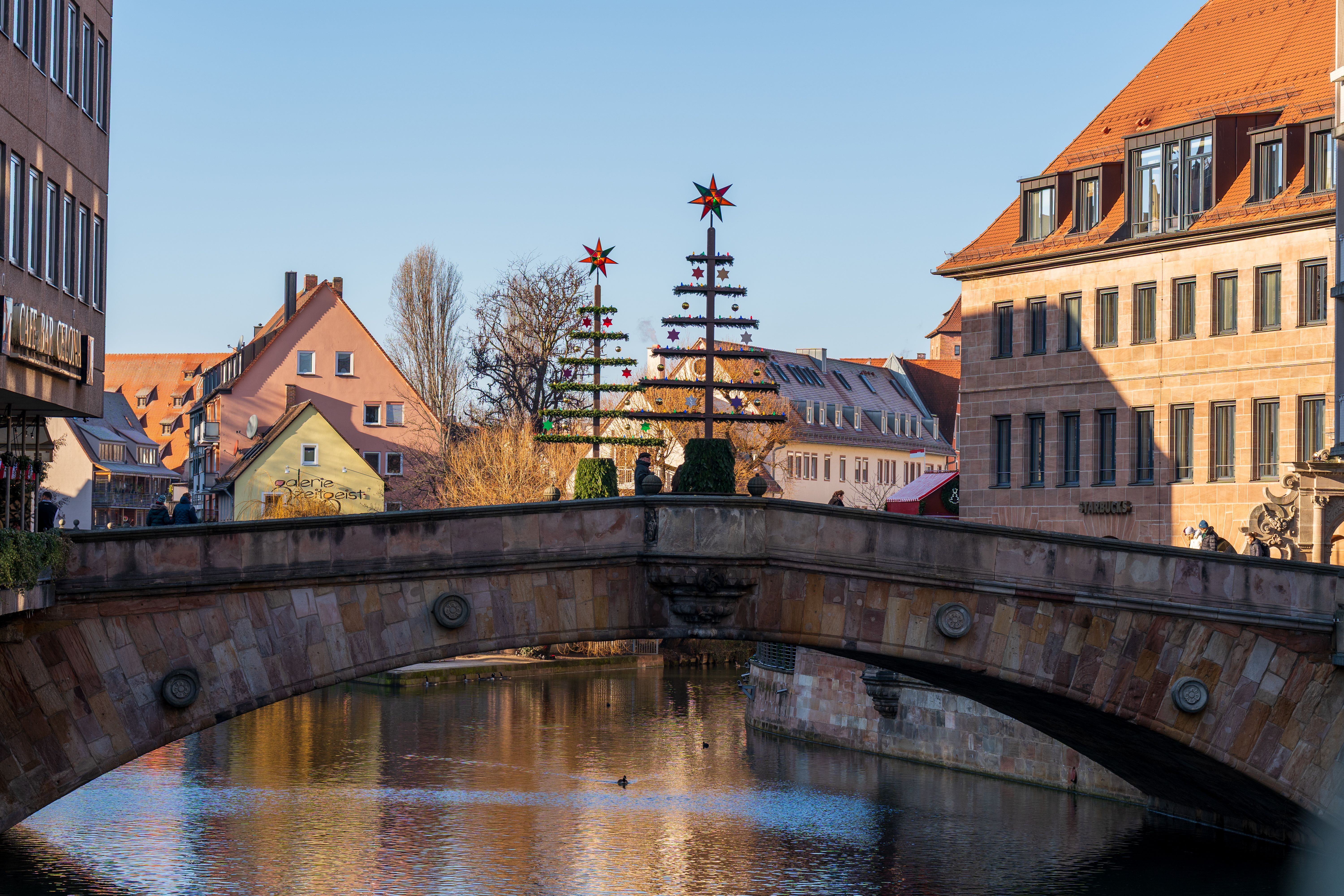 A stone bridge over a river with two tall decorated trees topped with red stars, and orange-roofed buildings behind under a blue sky