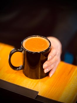 A cozy scene of a hand holding a black mug filled with hot coffee on a wooden table.