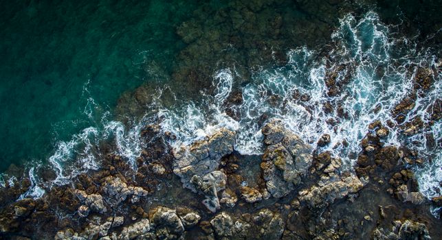 Drone shot capturing waves crashing against rugged rocks on a turquoise shoreline.