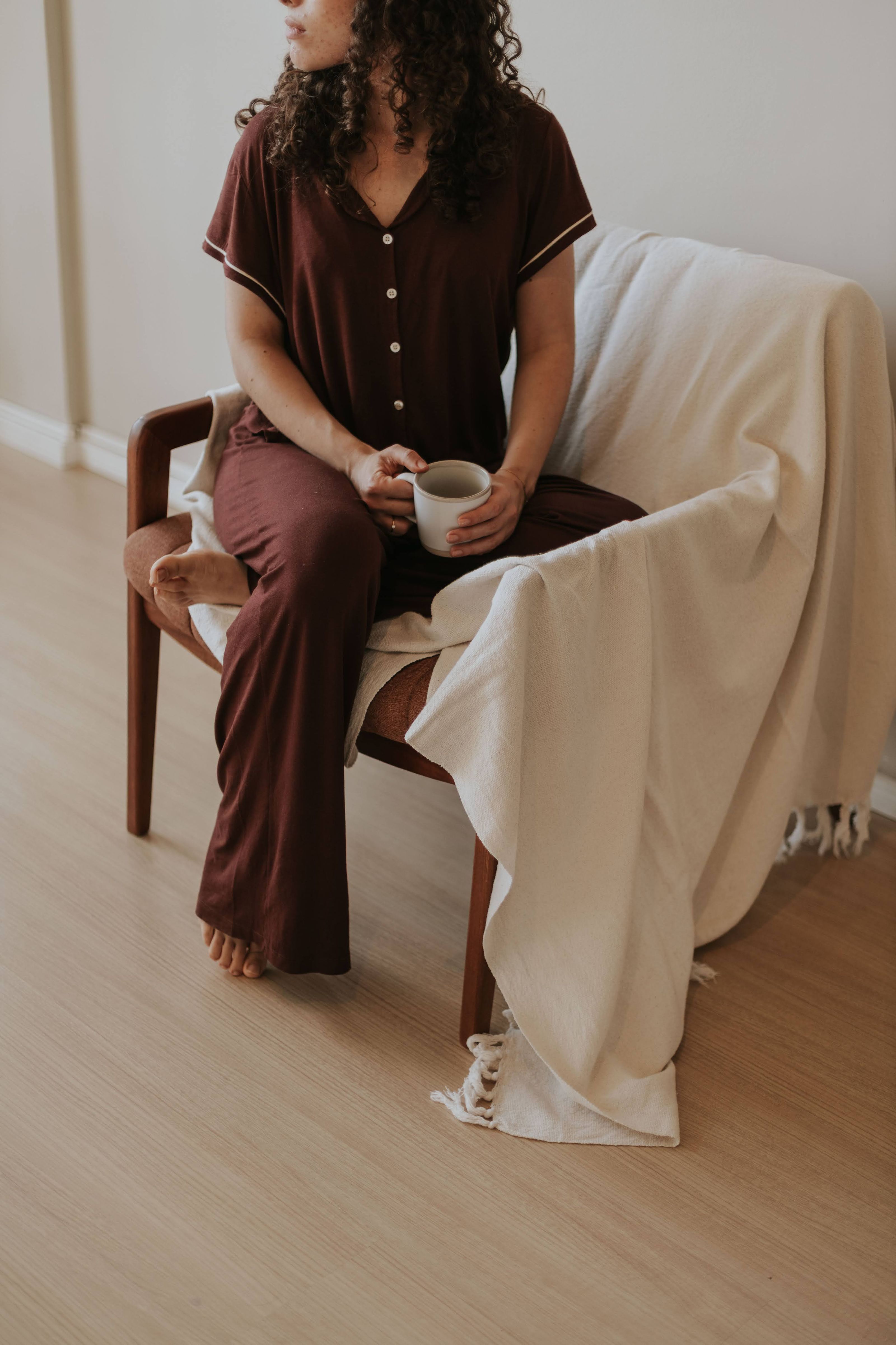 Mulher em pijama marrom desfrutando um café em uma cadeira aconchegante dentro de casa.