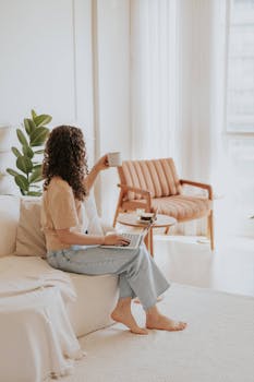 A woman enjoys a relaxing moment with coffee and a laptop in a bright modern living room setting.