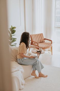 A woman sits relaxed on a couch using a laptop, embodying remote work and comfort in a modern interior.