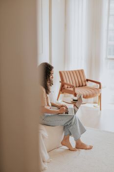 Young woman works on a laptop while sitting on a bed in a bright, cozy living room with a modern aesthetic.