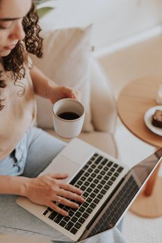 Woman enjoying coffee while using a laptop, sitting comfortably indoors.