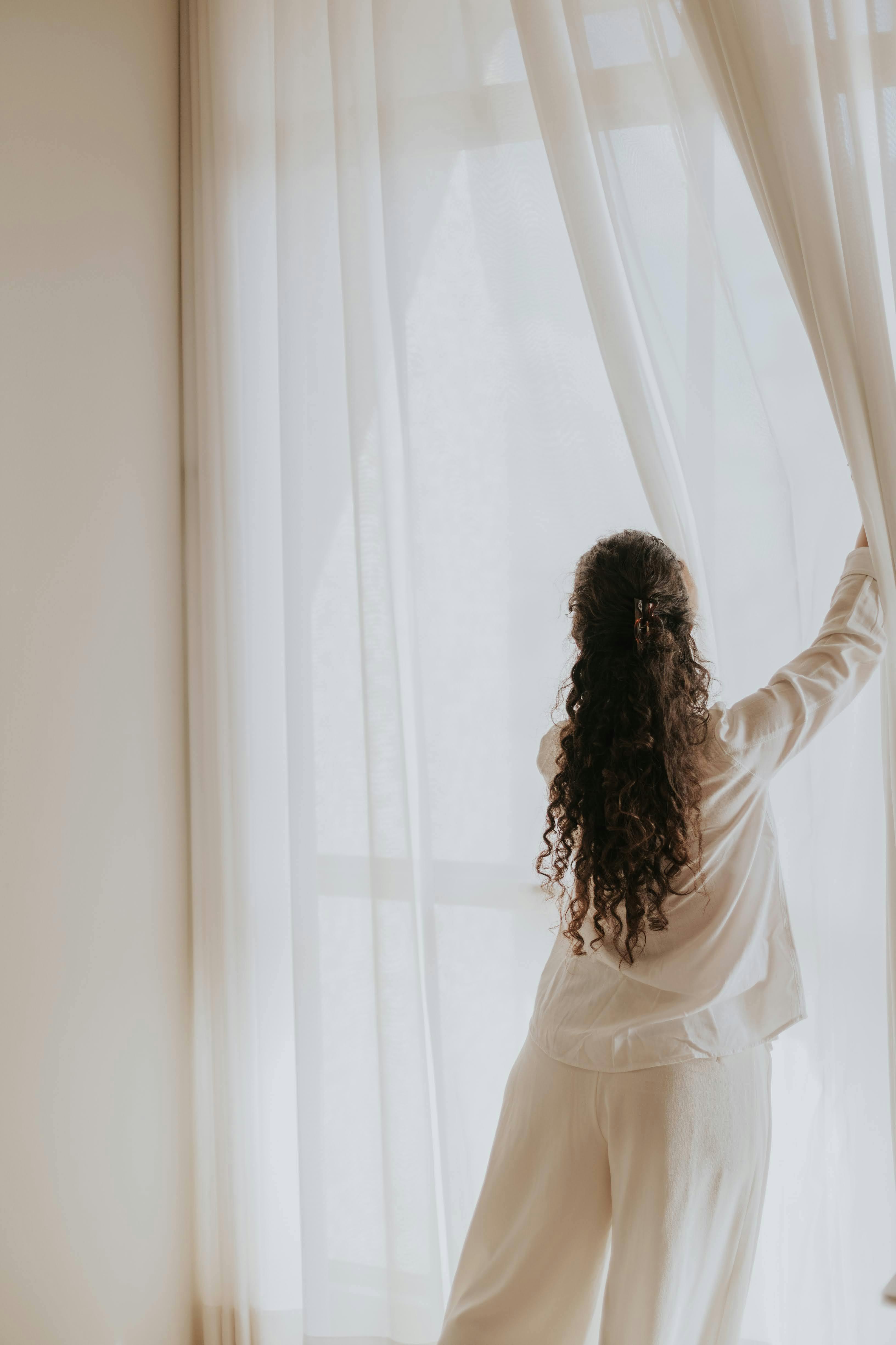 A woman opens curtains in a softly lit room, embracing the gentle daylight.