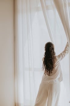 A woman opens curtains in a softly lit room, embracing the gentle daylight.