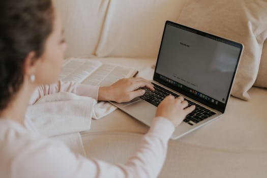Adult woman typing on a laptop with an open book nearby, working comfortably from her sofa.