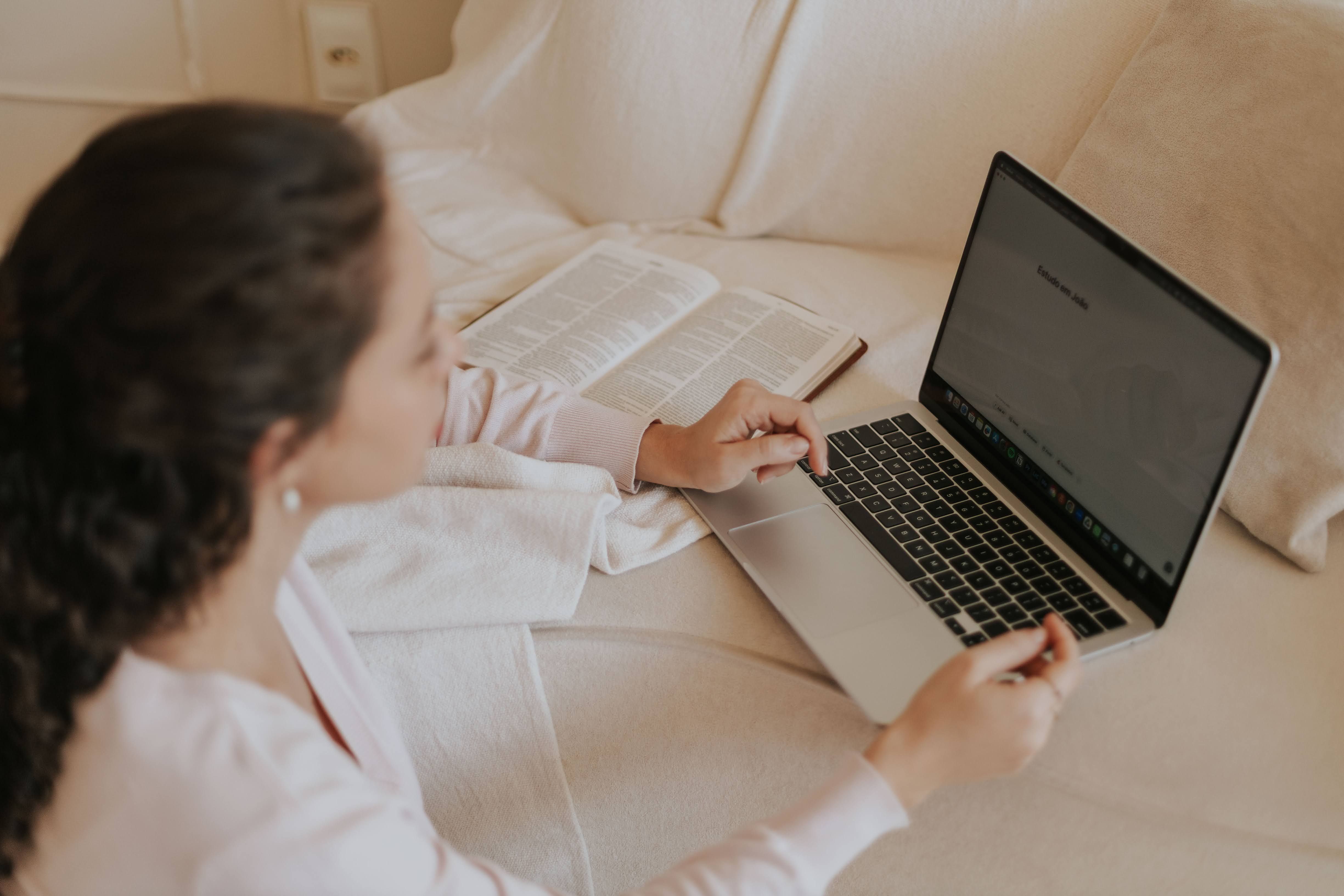 A woman reads a book and uses a laptop on a cozy sofa indoors, creating a comfortable study space.