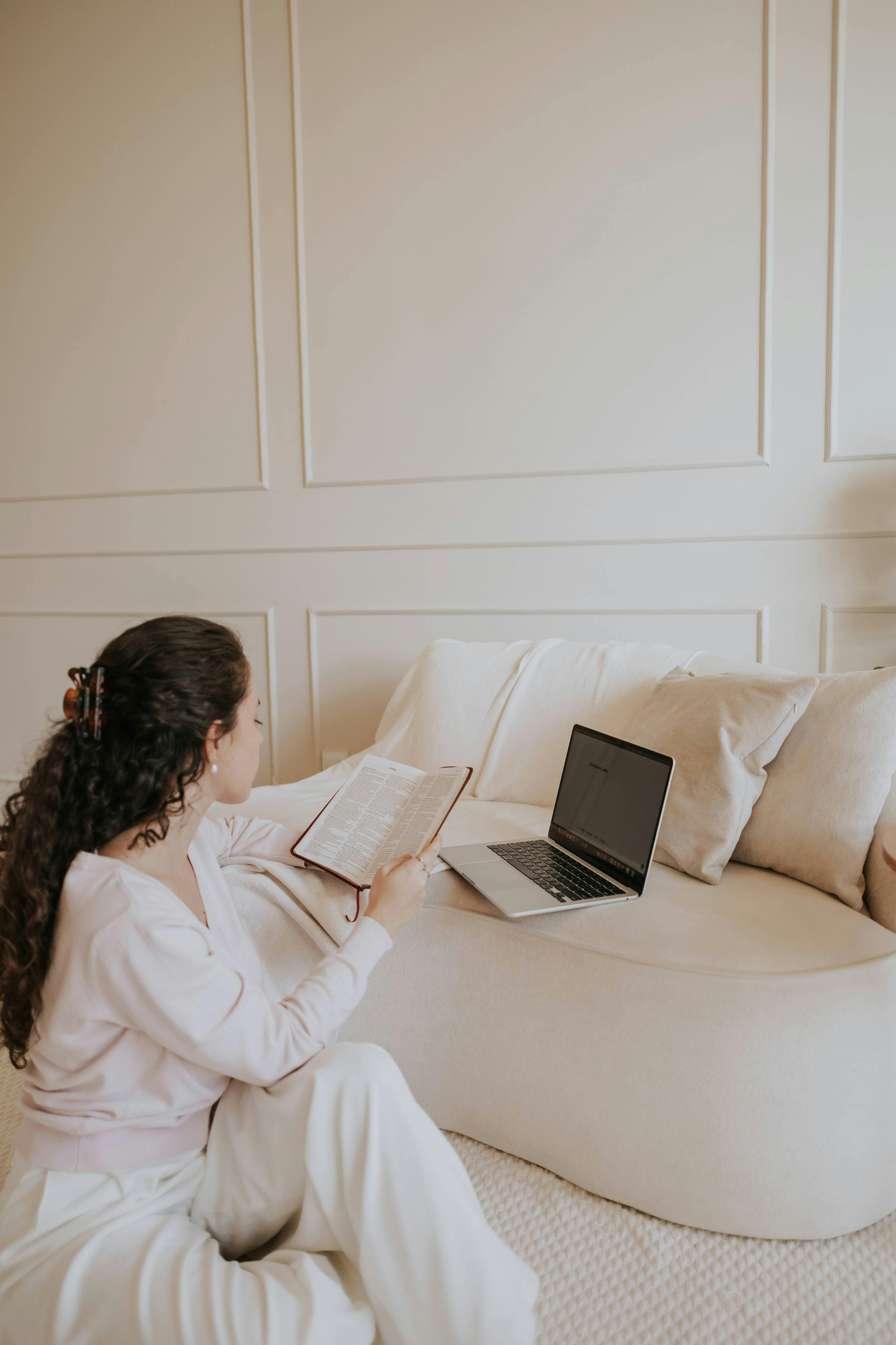 A woman reads a book in a cozy, minimalist room with a laptop on a sofa, embodying peaceful study vibes.