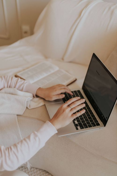 Free A cozy indoor setting with hands typing on a laptop, next to an open book on a soft surface. Stock Photo