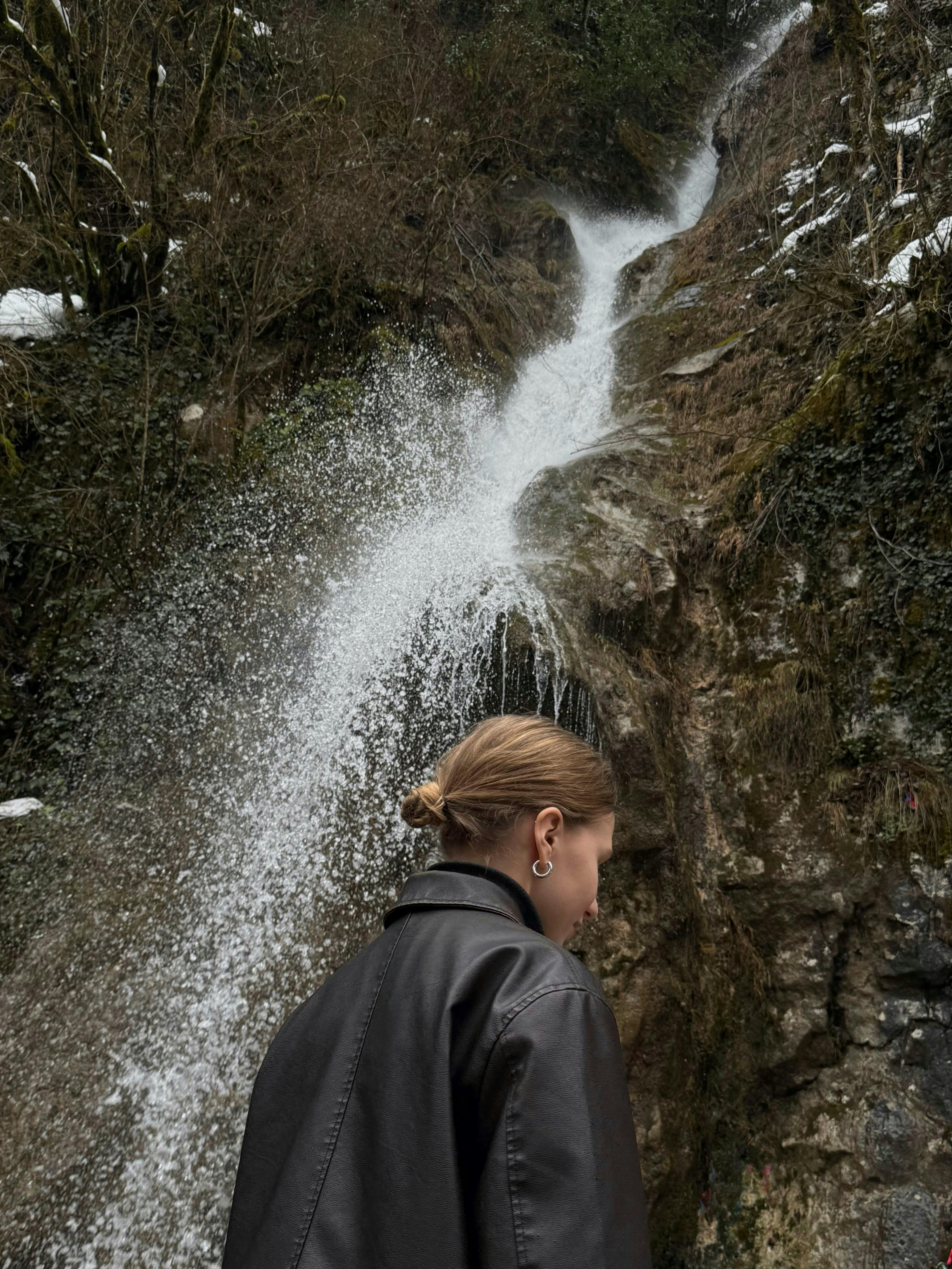 A woman in front of a cascading waterfall surrounded by lush greenery and rocky terrain.