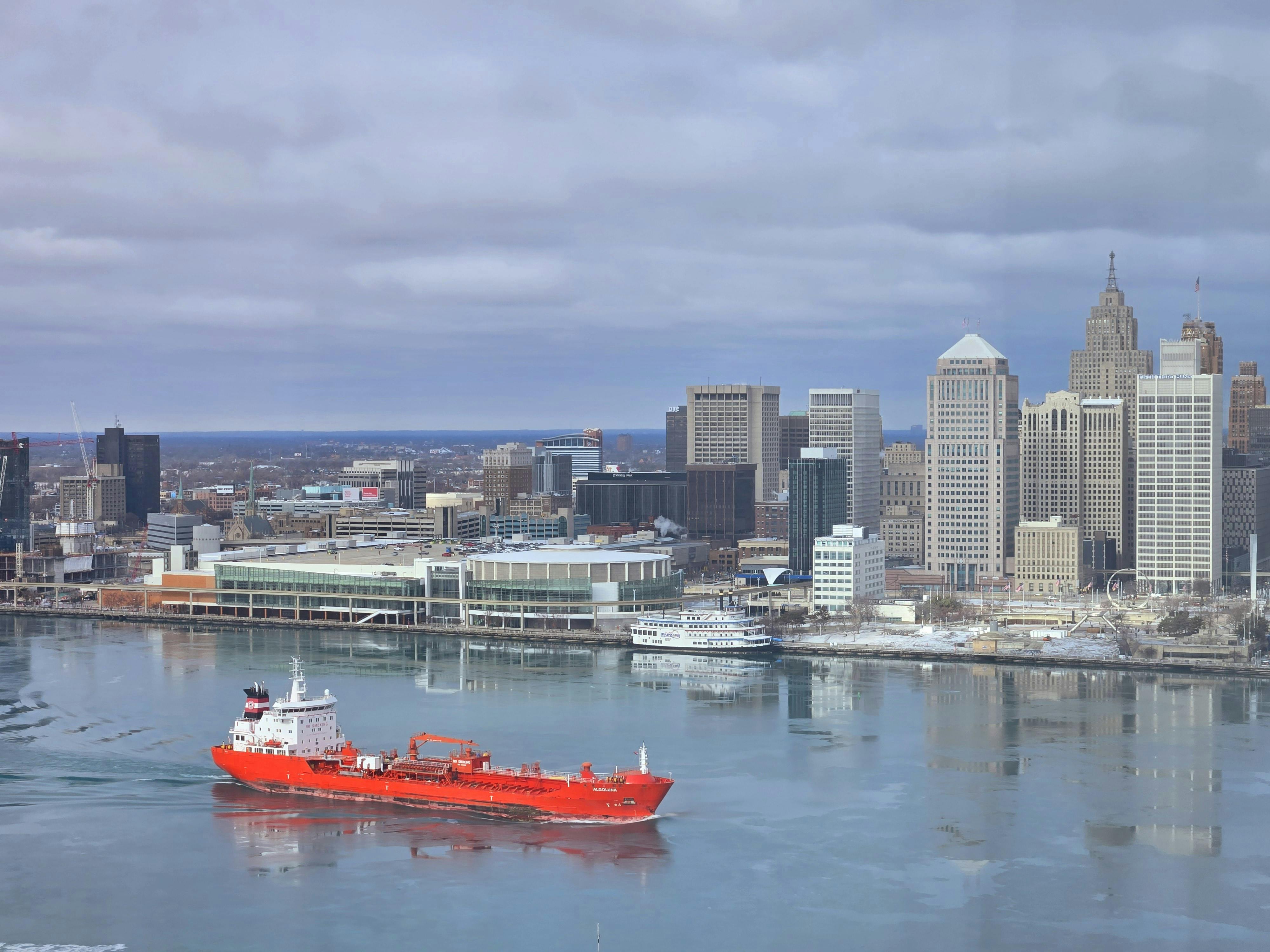 A vibrant red cargo ship sails through icy waters with the Detroit skyline in the background.