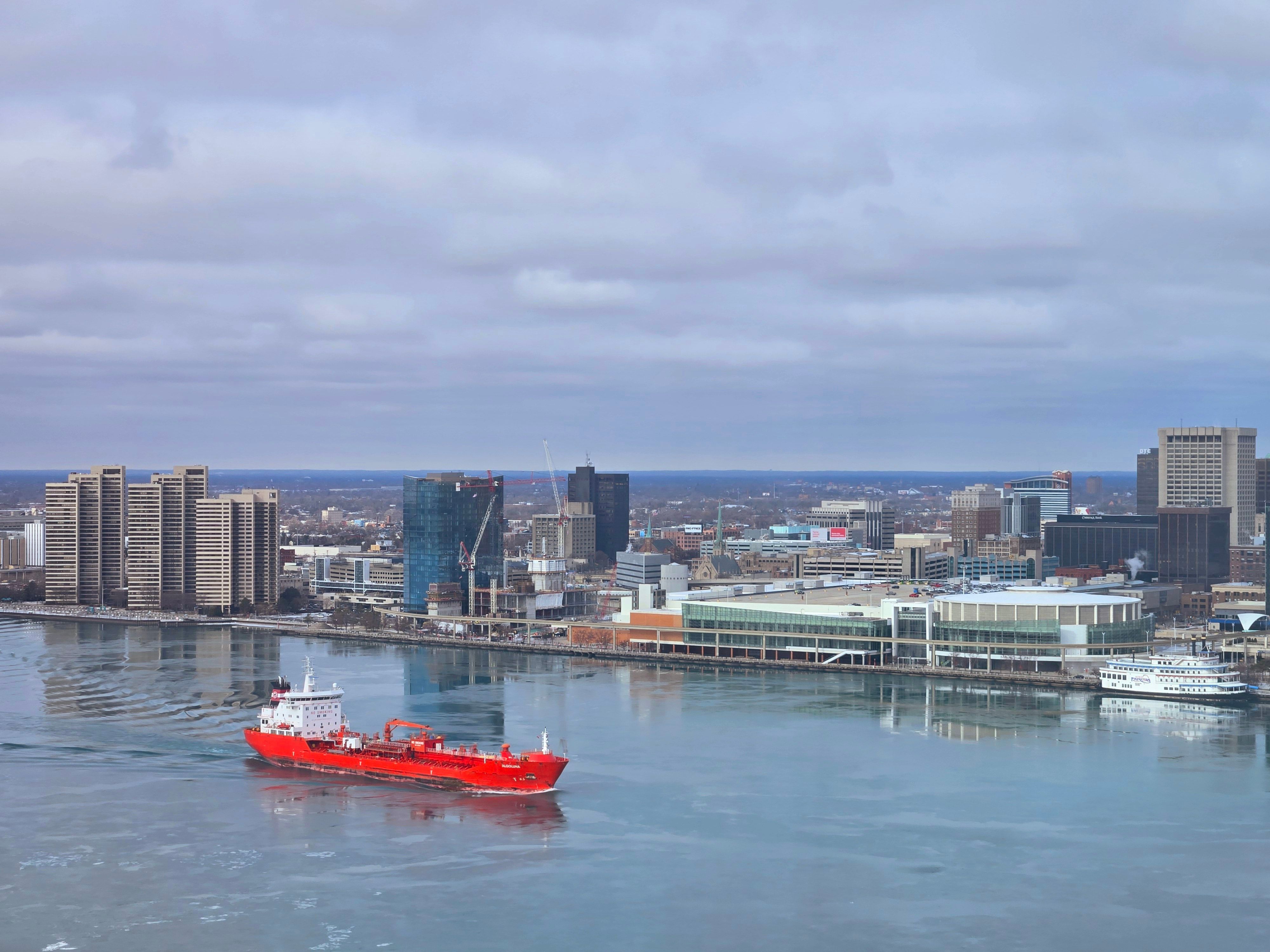 A red cargo ship sails on a calm river against a modern city skyline under a cloudy sky.