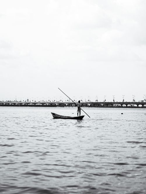 Free A fisherman rows a boat on calm waters near a prominent bridge under a cloudy sky. Stock Photo