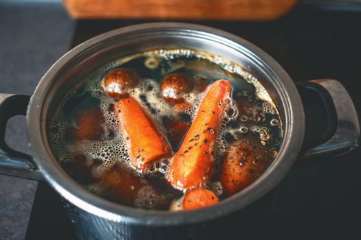 A close-up of carrots and potatoes boiling in a metal pot on a stove.