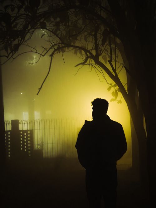 Free Mysterious silhouette of a person in a foggy park at night, with a glowing yellow light in the background. Stock Photo