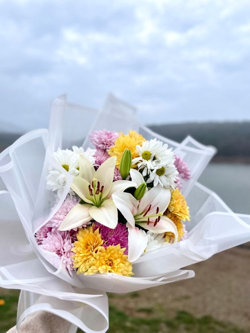 Free Colorful bouquet of lilies and chrysanthemums wrapped in white paper with a serene outdoor background. Stock Photo