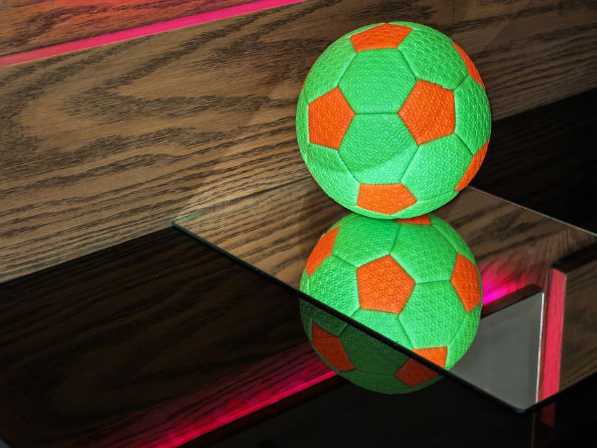 A green and orange soccer ball reflects on a shiny glass surface with a wooden background.