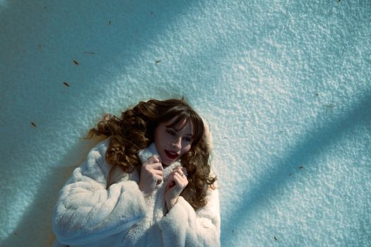 A woman in a winter fur coat lies on the snow, enjoying the cold with a smile on her face.