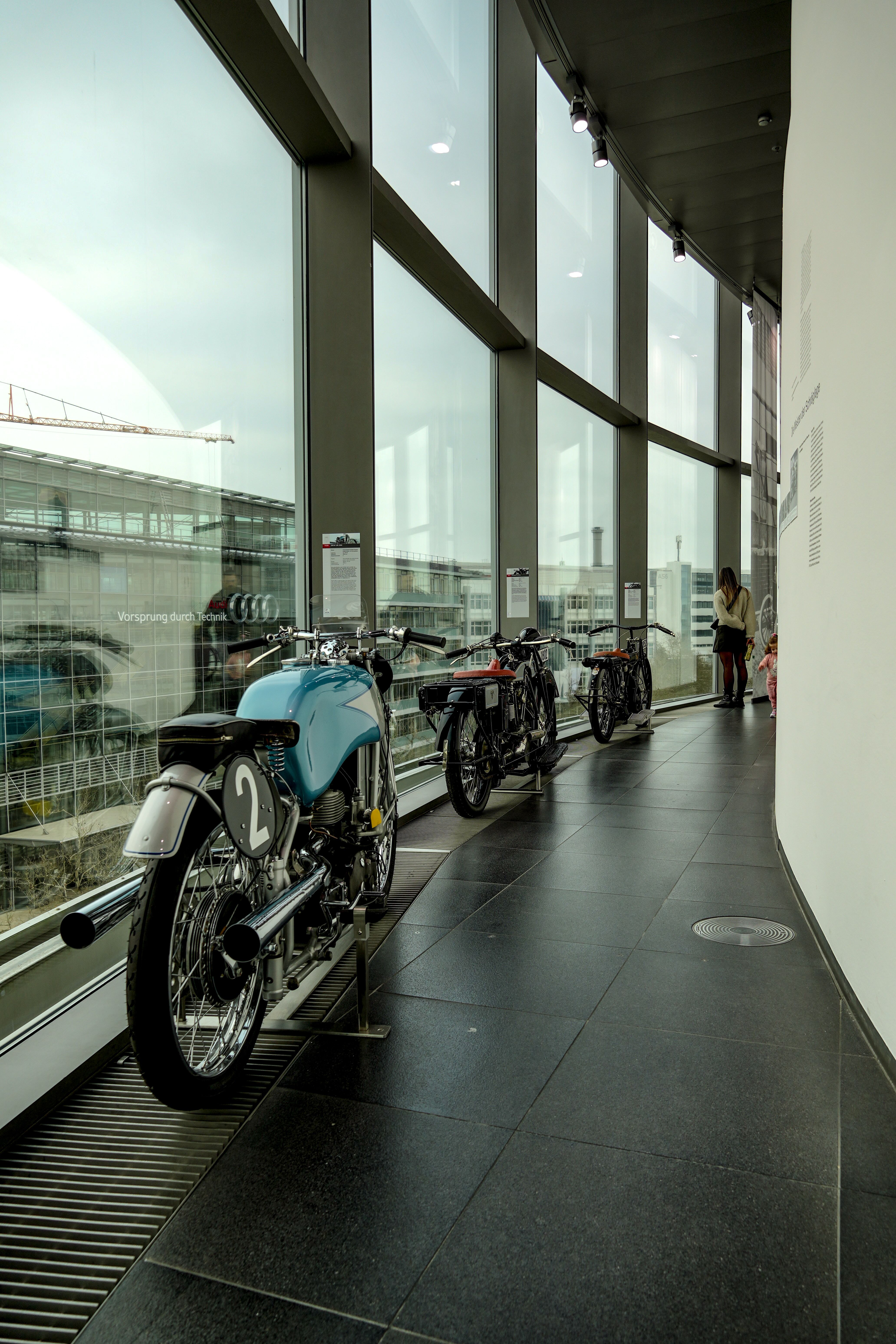 Vintage motorcycles displayed in a modern German museum overlooking the cityscape through large windows.