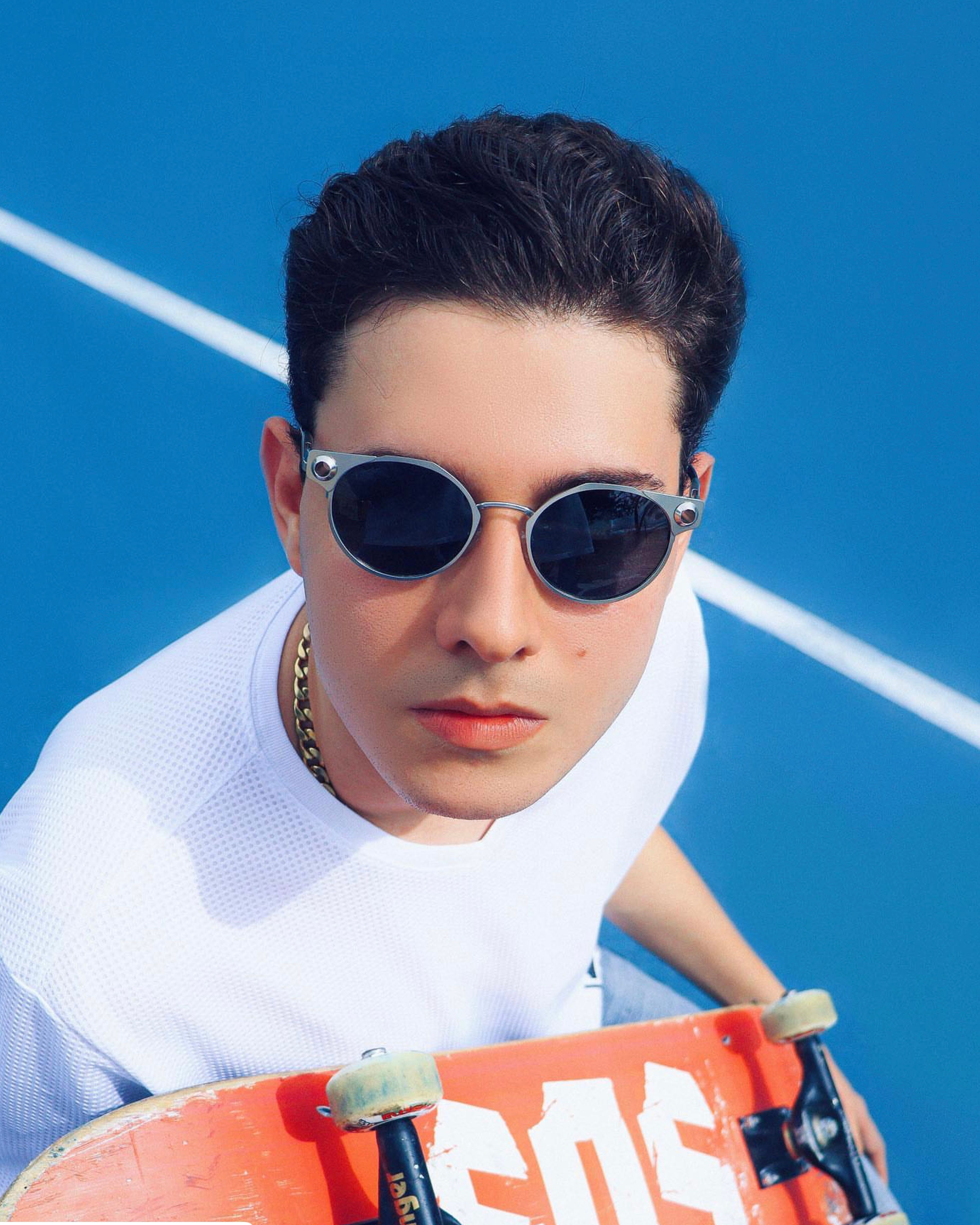 Trendy young man in sunglasses holding a skateboard on a vibrant blue sports court.