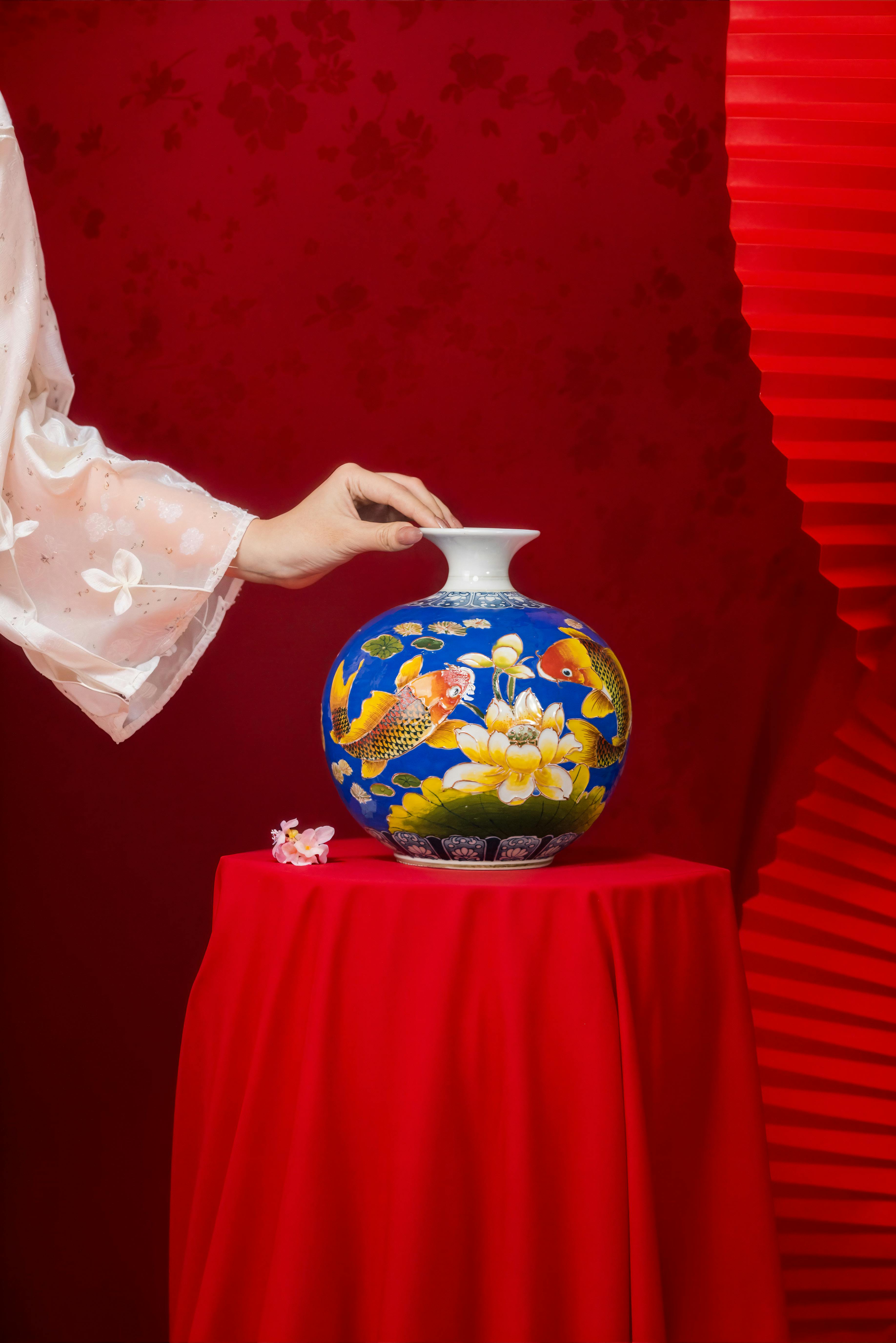 Δωρεάν Ένα χαριτωμένο χέρι τοποθετεί ένα λεπτομερές μπλε βάζο στολισμένο με ψάρια koi και λουλούδια σε ένα ζωηρό κόκκινο φόντο. Φωτογραφία από στοκ φωτογραφιών