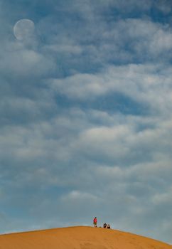 A group of people stand atop a sand dune under a partly cloudy sky with a visible moon.