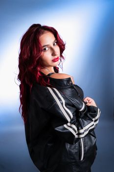 Confident woman with red hair wearing a leather jacket poses in a studio with blue lighting.