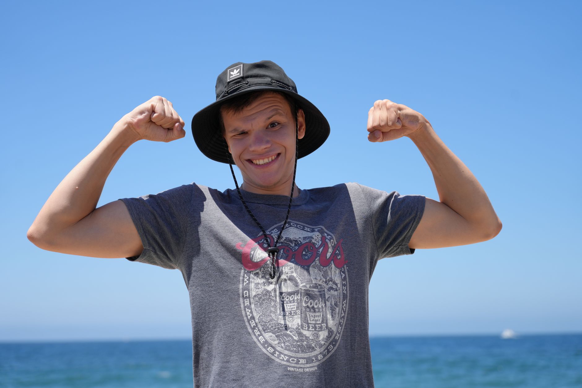 Smiling man wearing a hat and t-shirt flexing muscles on a sunny beach day.