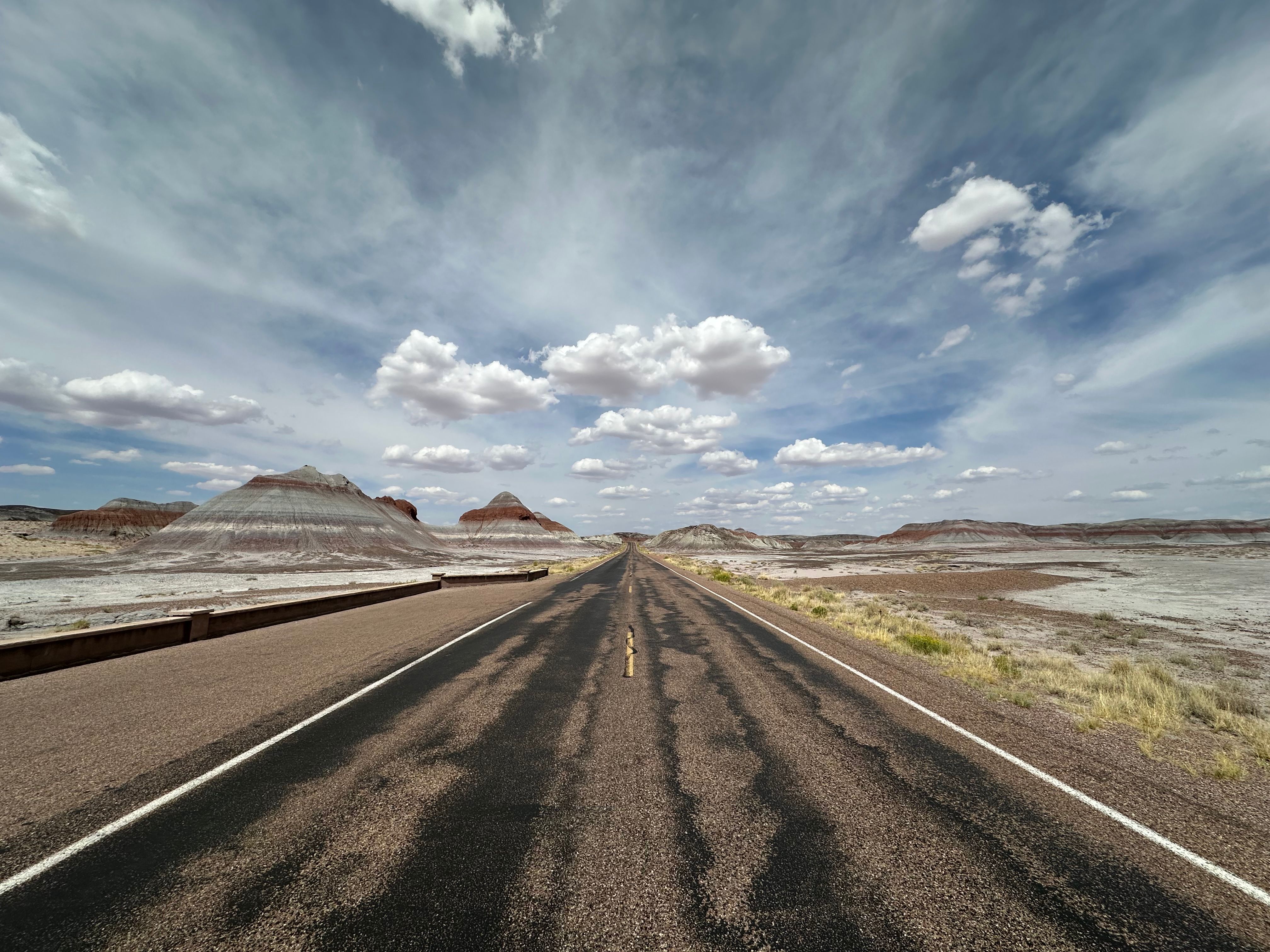 Paved road through Arizona's Painted Desert with striking rock formations and a vast, cloudy sky.