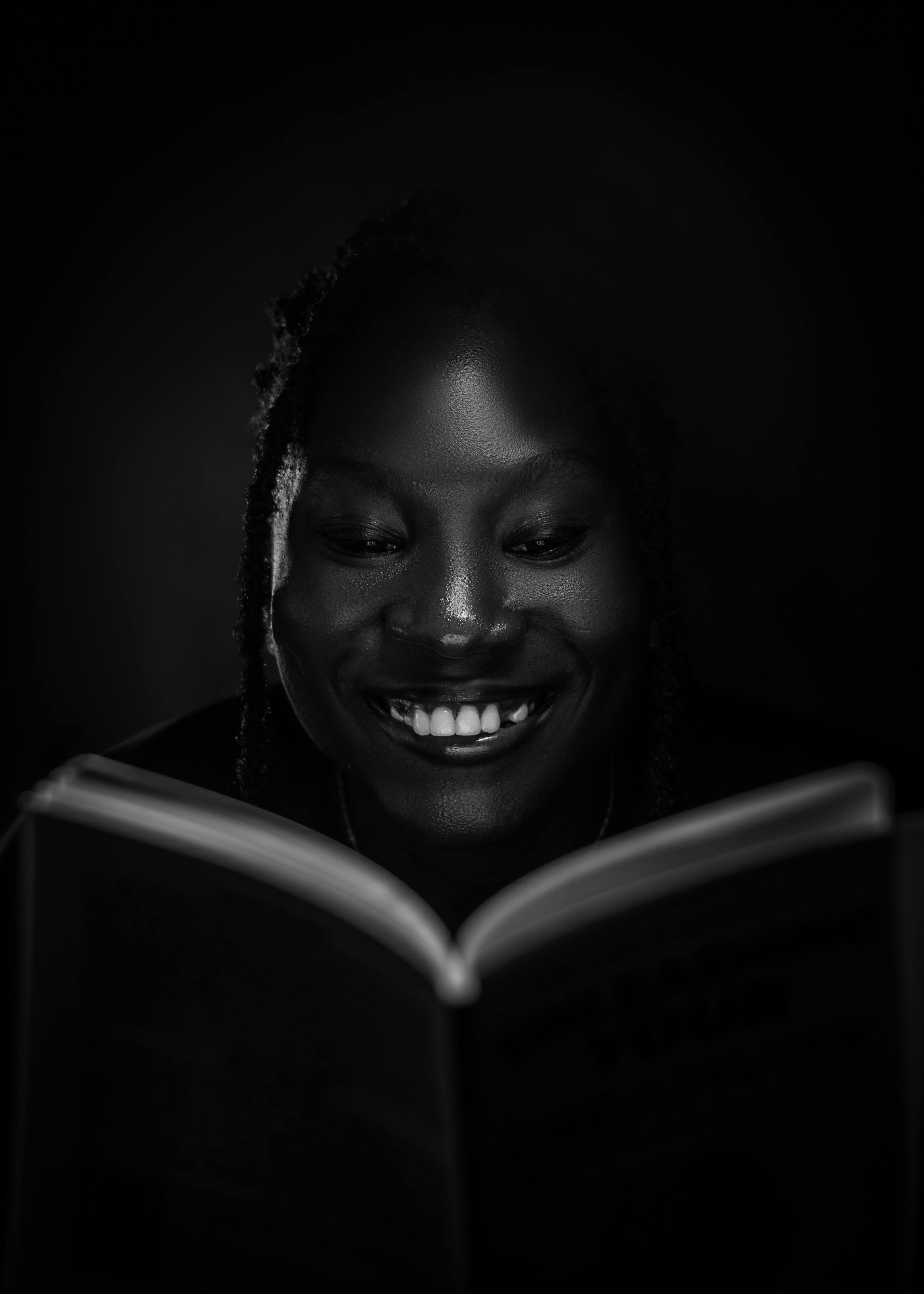 Gratis Una donna legge allegramente un libro in una stanza scarsamente illuminata, creando un'atmosfera serena e concentrata. Foto a disposizione