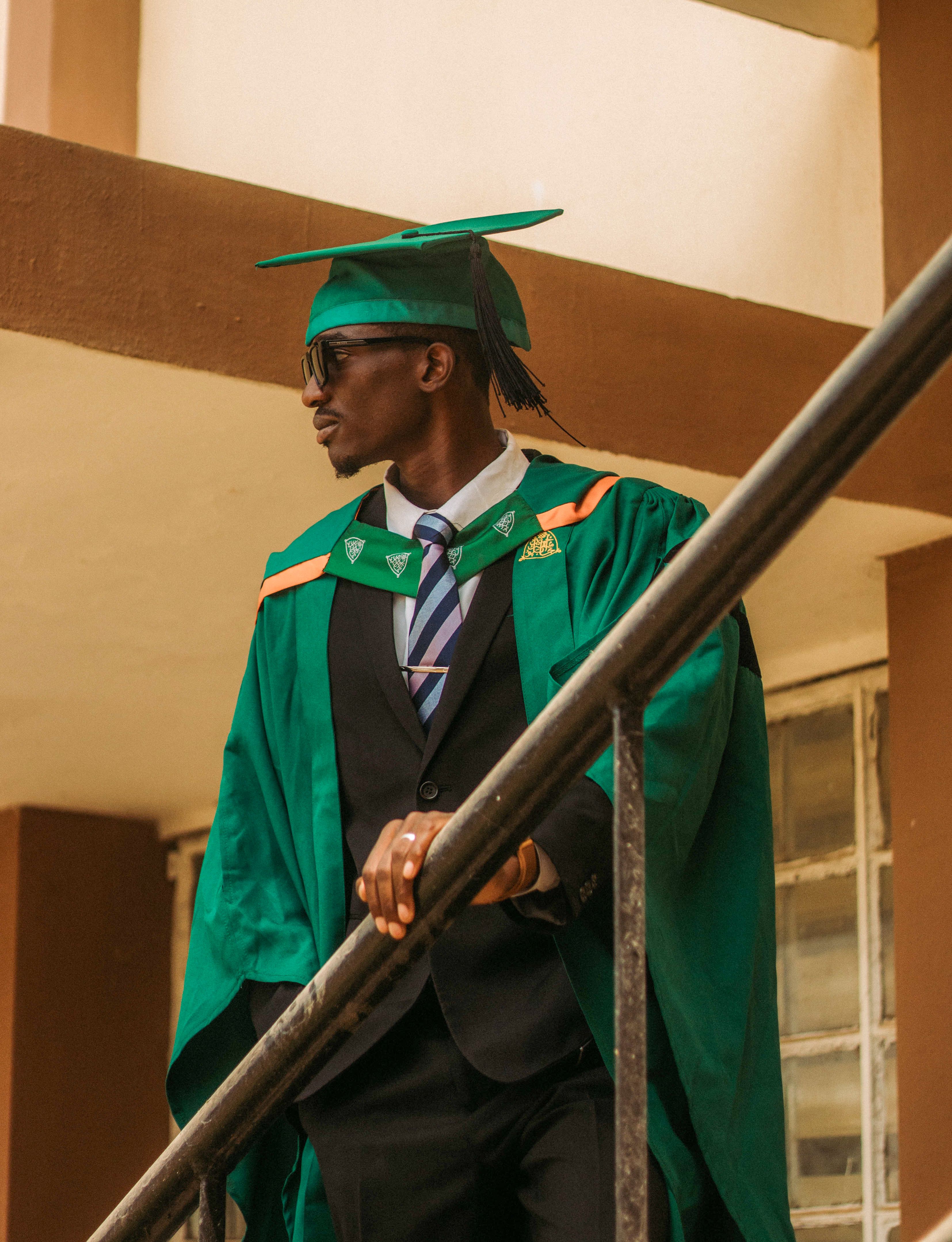 Confident young graduate in green cap and gown standing on staircase with sunglasses.