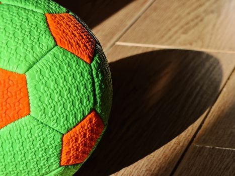 Bright green and orange soccer ball casting shadow on sunlit wooden floor.