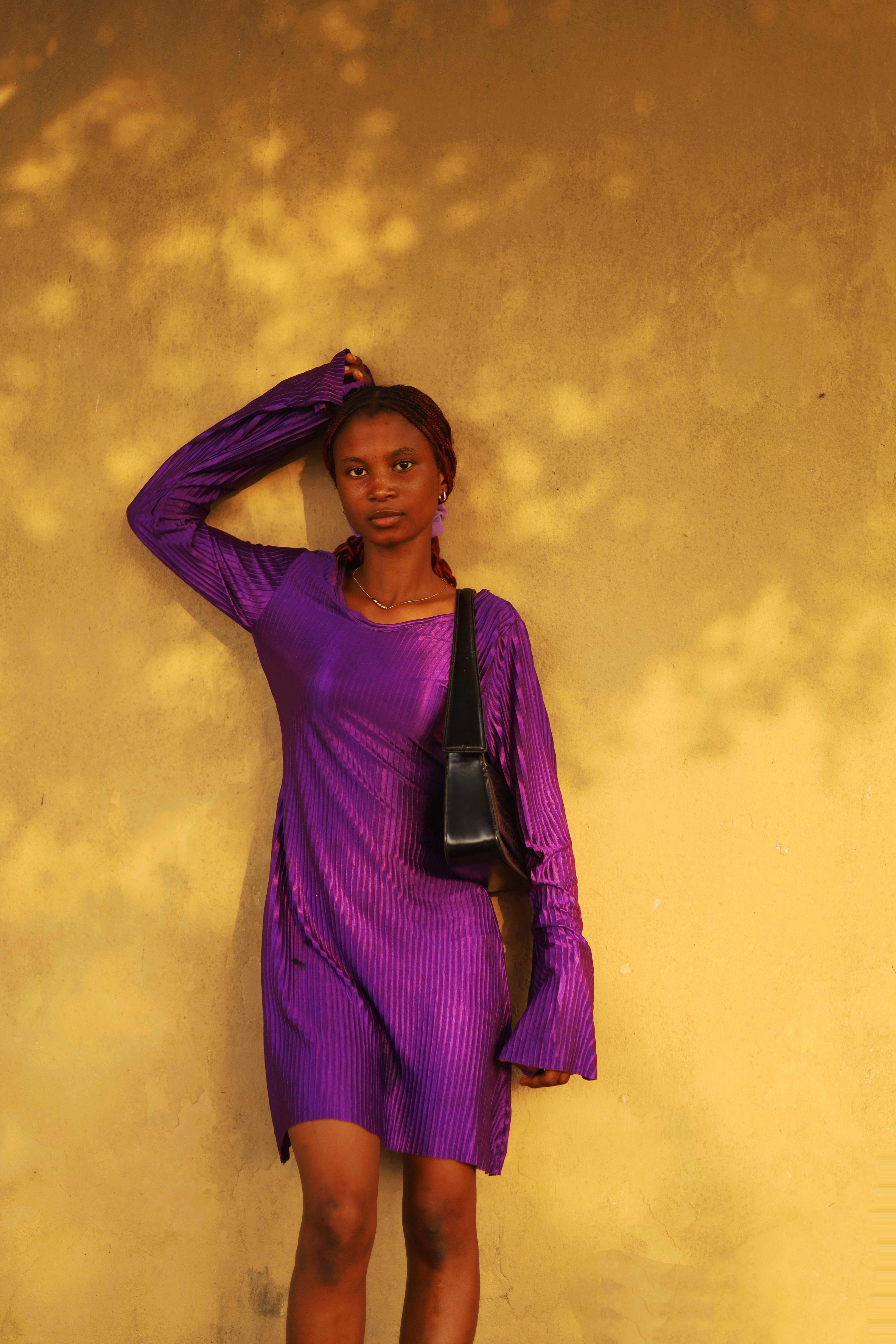 grátis Mulher elegante com um vibrante vestido roxo em pé contra uma parede iluminada pelo sol. Foto profissional