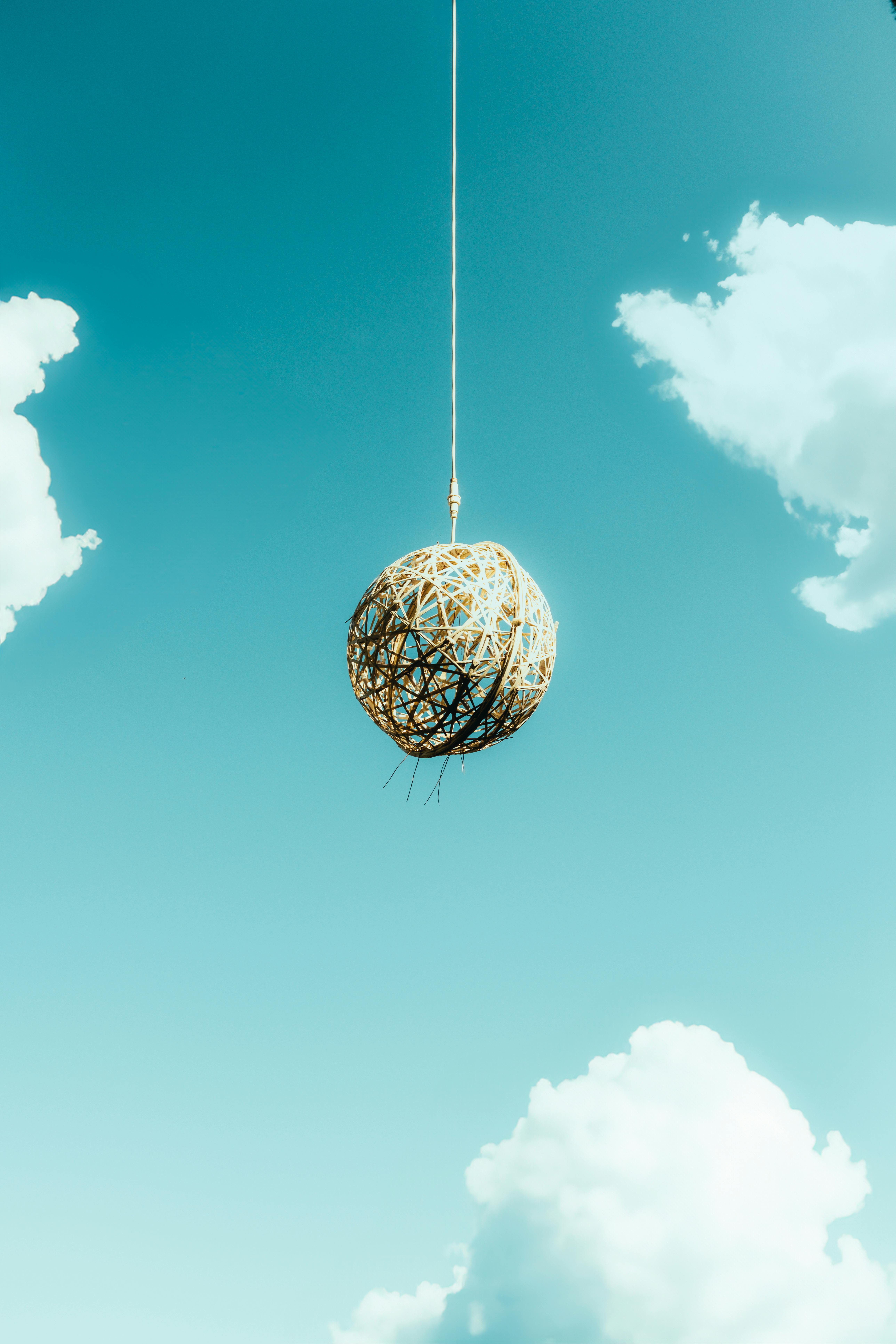 免费 一个藤编球悬挂在晴朗的蓝天和朵朵白云之间，营造出一种宁静而抽象的氛围。 素材图片