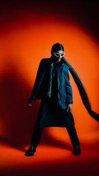Fashion-forward individual in dark attire poses dynamically with sunglasses and flowing scarf.
